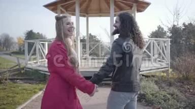积极的两族青年夫妇站在秋天公园的亭子前，笑着说话。 快乐的嬉皮士